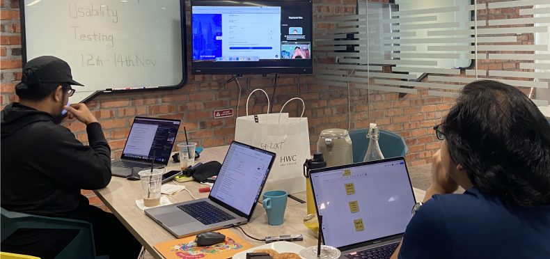 A usability testing session being observed from observer room. Features a pair of researcher note-taking the session using their laptop.