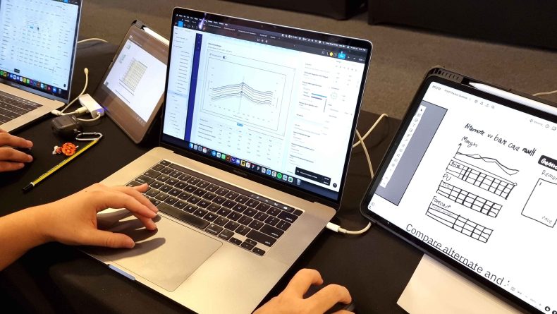 A close-up of designer’s hands interacting with a laptop displaying a prototype of a data visualization dashboard. Another iPad screen on the right shows a digital sketch of the data dashboard for design reference,