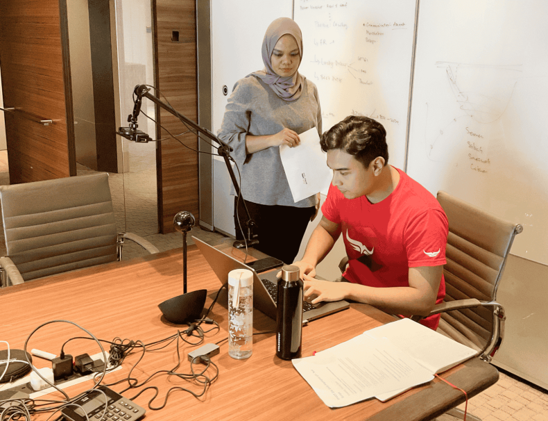 A man in a red shirt is seated at a conference table, setting up a usability testing rig on a laptop, with a microphone and various cables connected. A woman in a hijab stands nearby holding a notepad, reviewing notes. The setting is a professional meeting room, prepared for a live usability test with an enterprise leadership team.